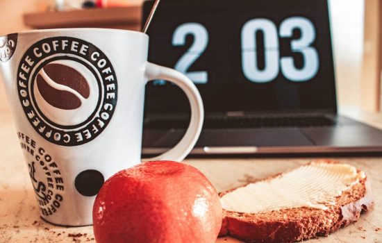 desk with a coffeecup slice of bread, a mandarine and a laptop with clock